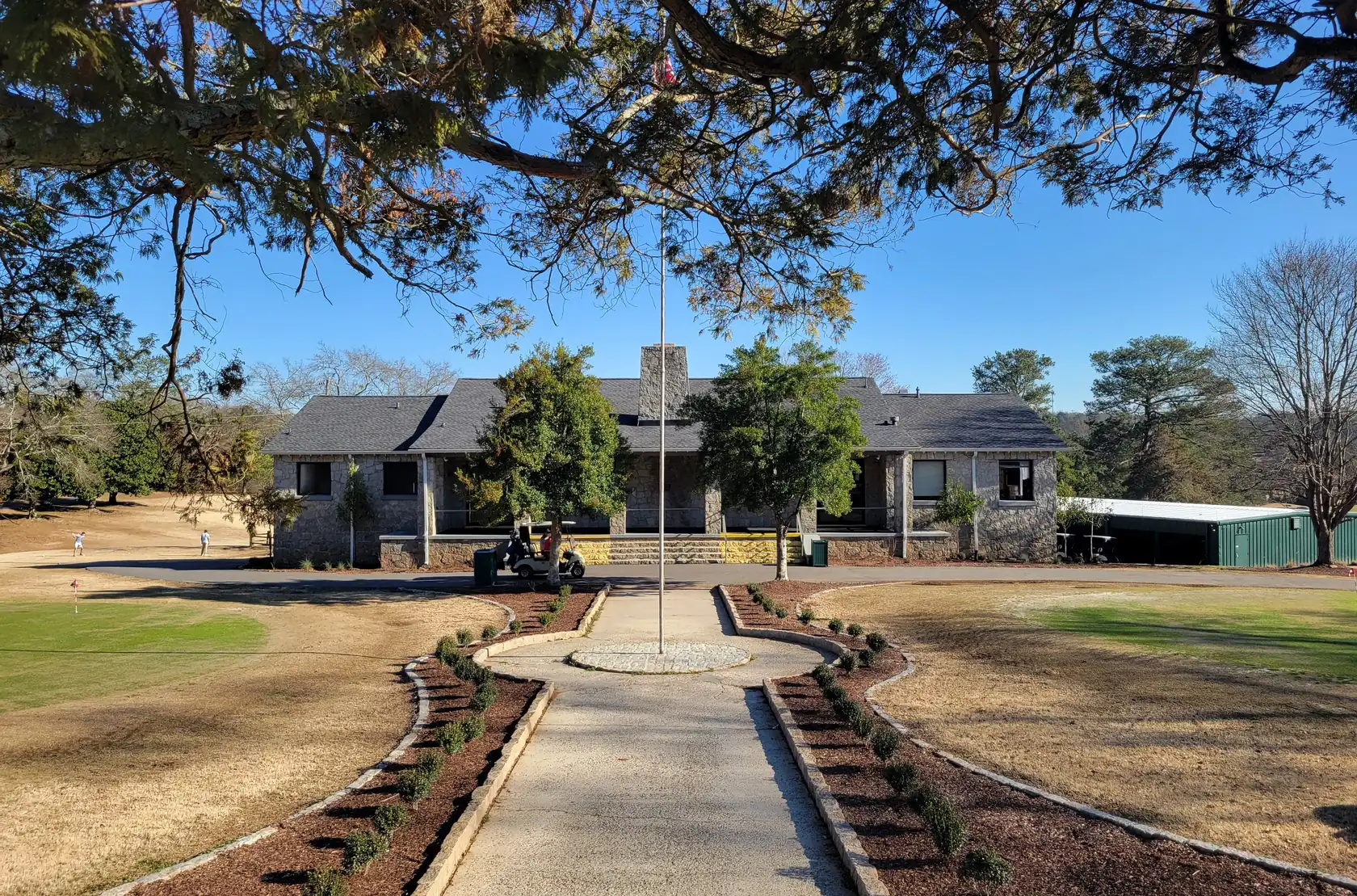 A straight walk between mulched beds leading to a low stone clubhouse with a dark shingle roof and a deep porch, golf carts by the steps and a putting green at left.