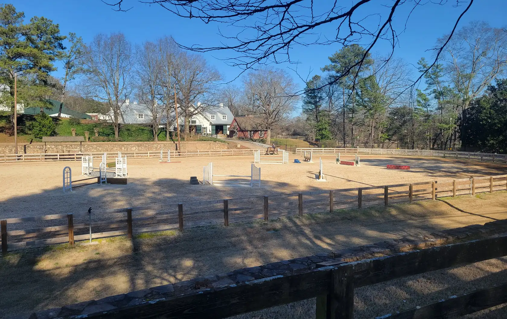 A sand riding arena set with white show jumps behind a split-rail fence and low stone wall, white stable buildings and a small red barn beyond, under a clear blue sky.