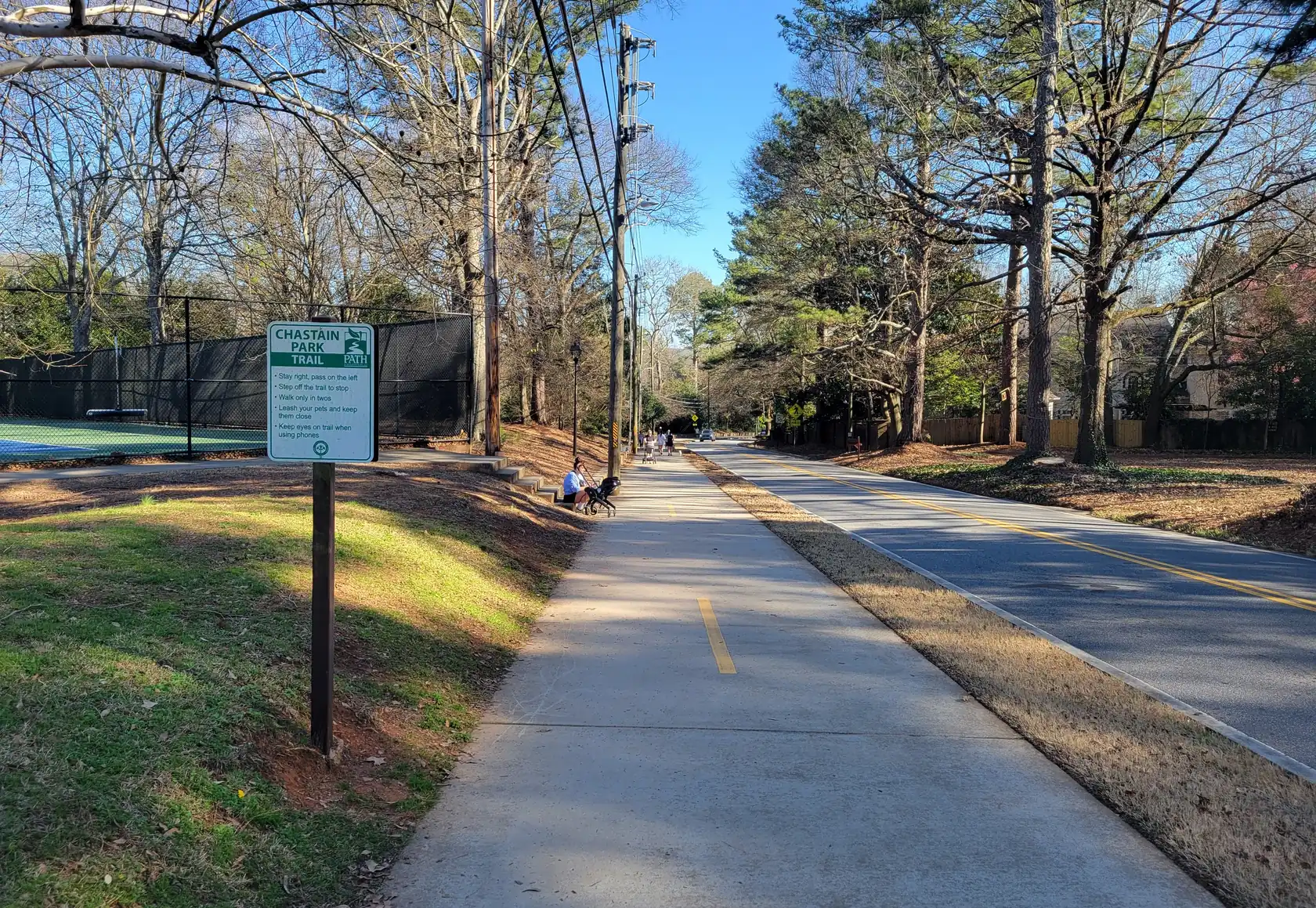 A wide concrete multi-use path with a yellow centre stripe running beside a two-lane road under tall pines and bare hardwoods, a Chastain Park Trail sign at left.