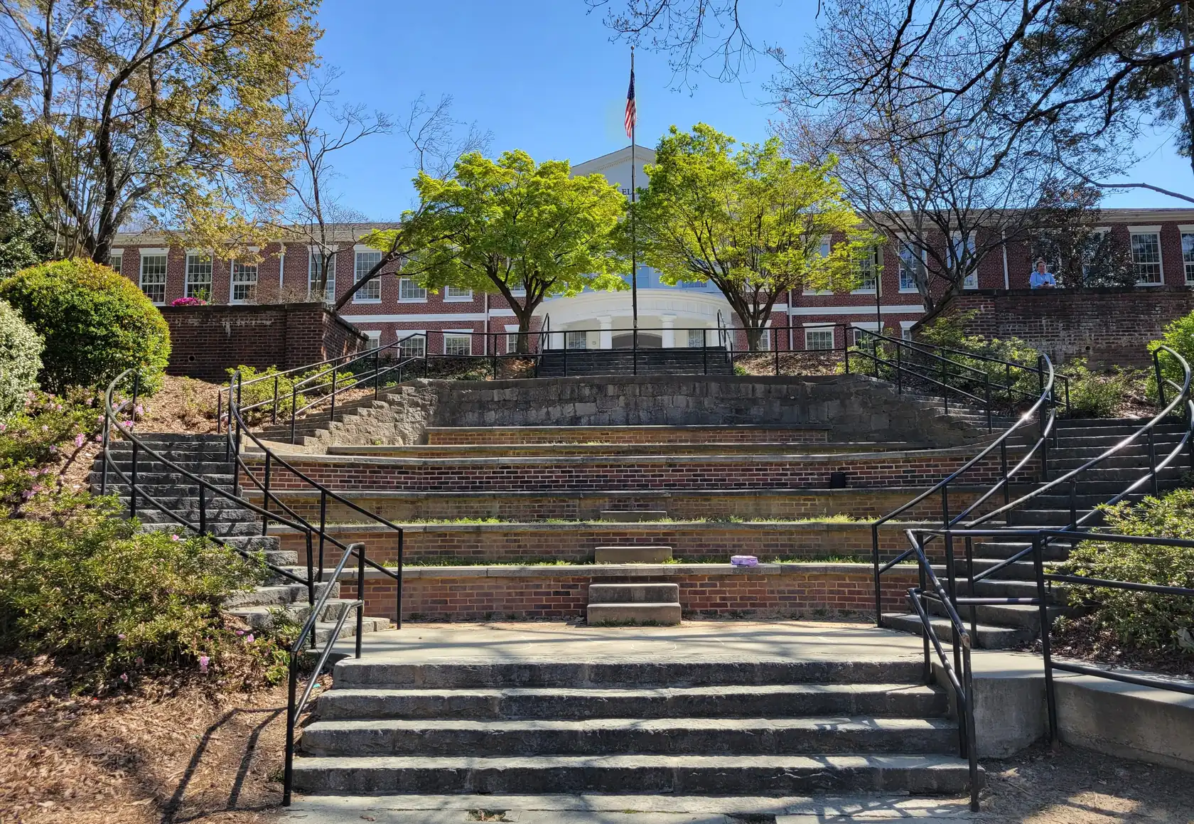 Curved granite and brick tiered steps rising through a small terraced amphitheater to a long red-brick building with a white columned entrance, pink azaleas in bloom at left.