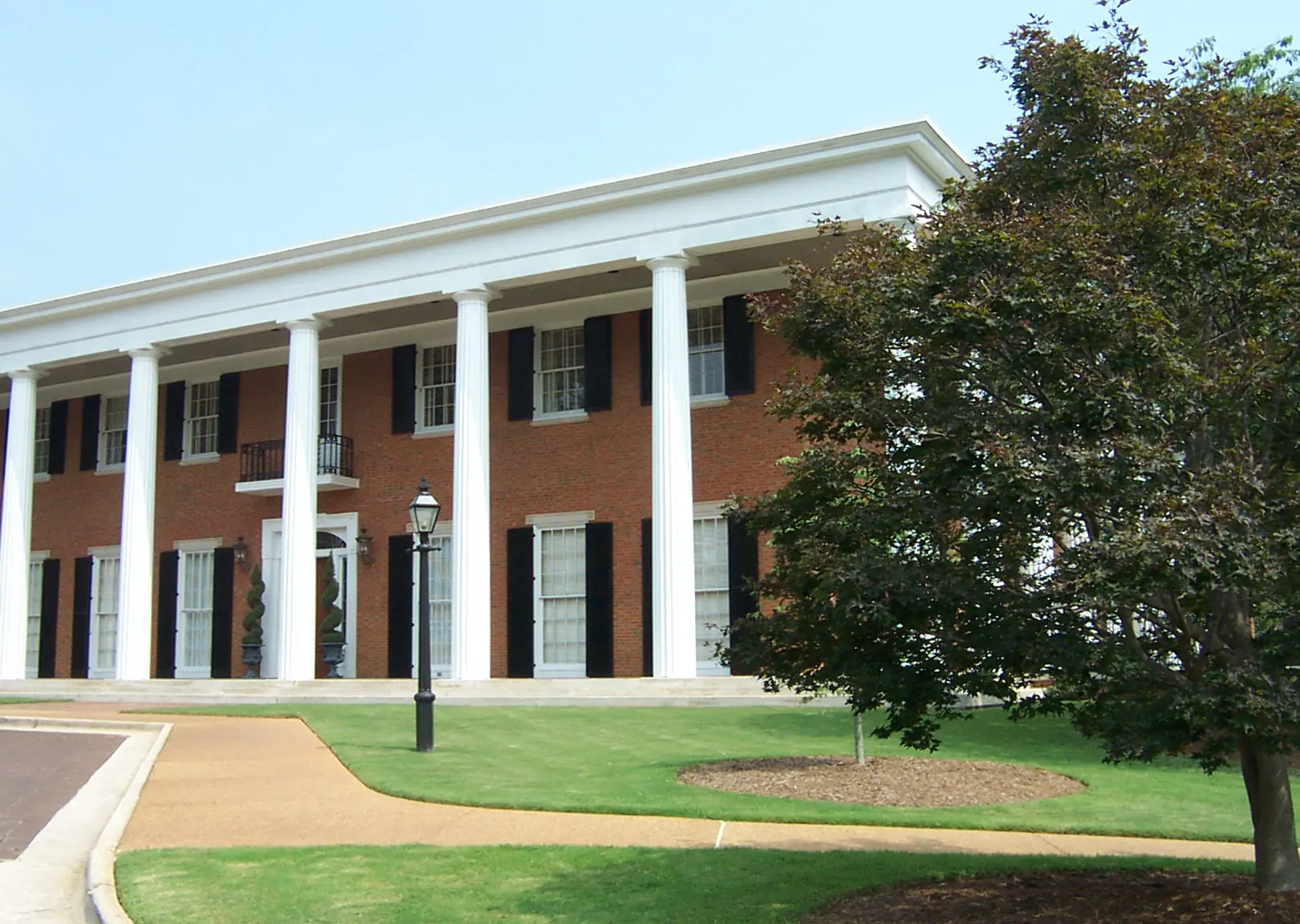 A red-brick Greek Revival mansion with tall fluted white columns, black shutters and a white entry door flanked by topiary urns, behind a manicured lawn and a lamp post.