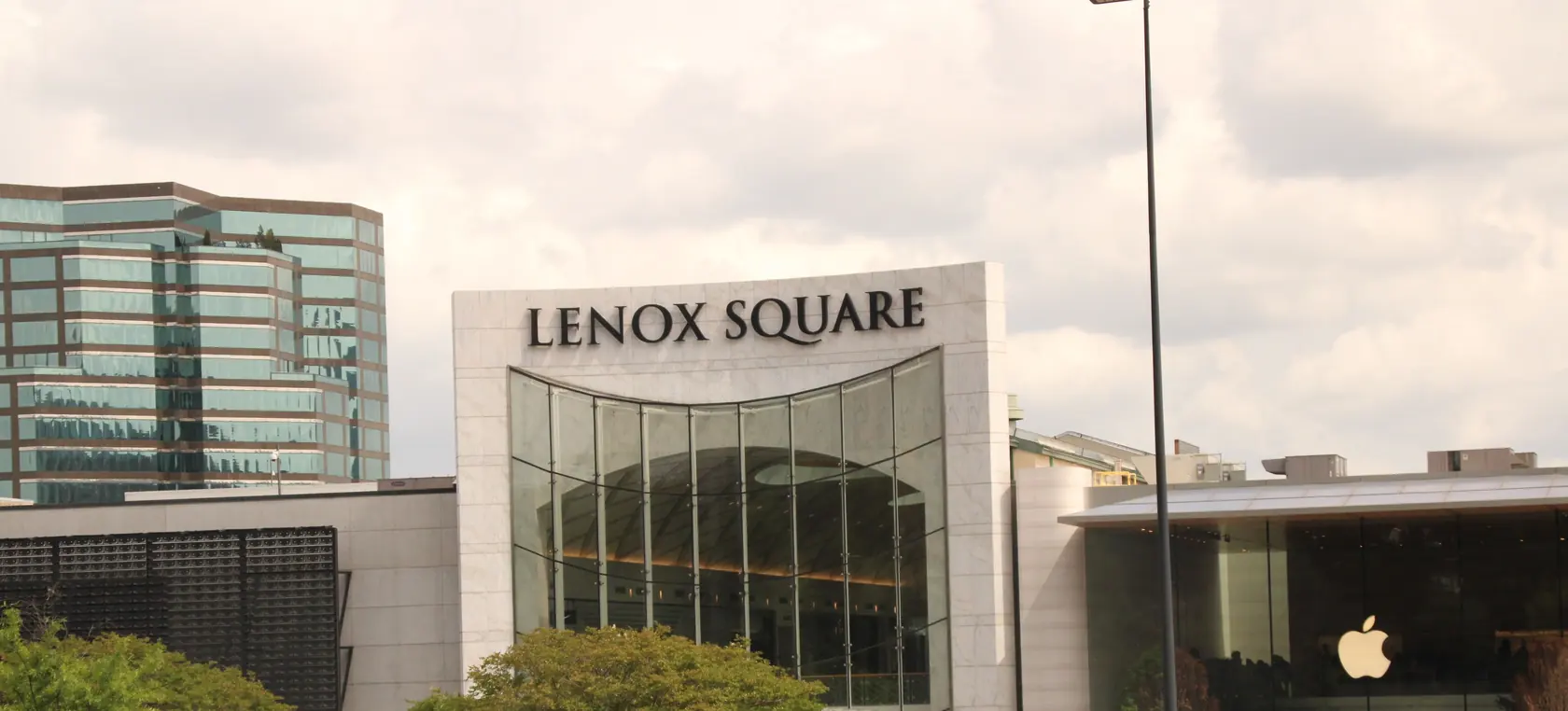 A tall pale stone frame lettered Lenox Square around a curved glass entrance wall, with a stepped green-glass office tower at left and young street trees in front.