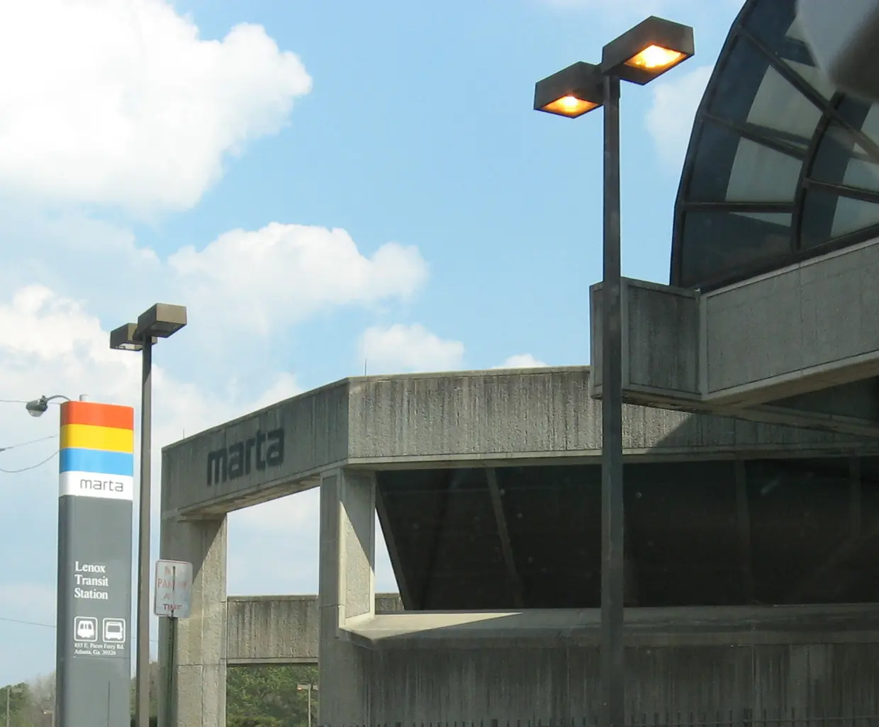 A board-formed concrete transit station with marta lettered on a curved canopy band, lamp posts and a striped MARTA pylon reading Lenox Transit Station, under a blue sky.