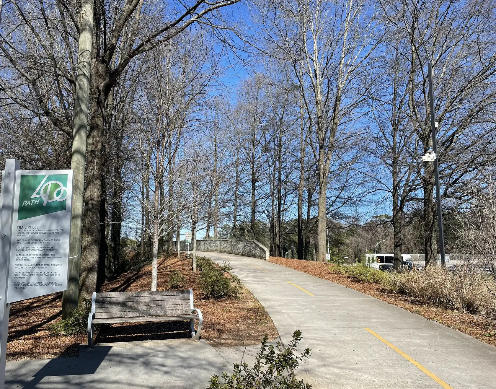 A wide concrete trail with a yellow centre line curving up to a low stone-walled bridge between tall bare trees, under a clear blue late-winter sky.