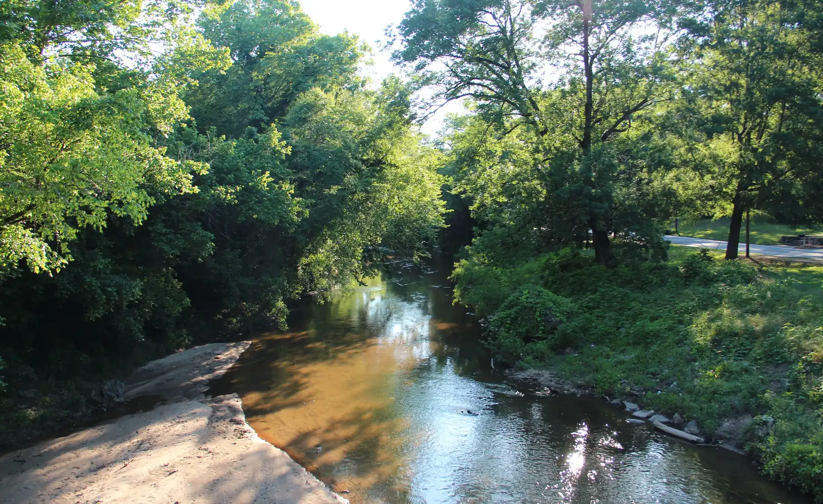 A shallow tea-coloured creek reflecting low sun as it runs between dense leafy banks, a pale sandbar on the left and a mown lawn and path on the right.