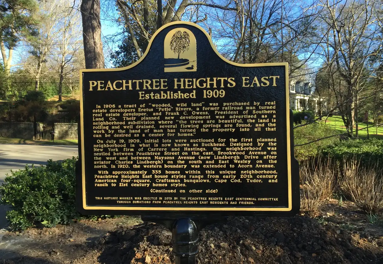 A black-and-gold cast historical marker headed Peachtree Heights East, Established 1909, in front of a curving street, a stone wall with an iron gate and bare winter trees.