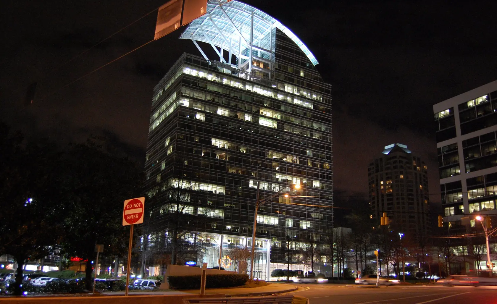 A lit glass office tower crowned by a curved illuminated canopy at night, a round-capped residential tower glowing behind it and light trails from traffic on the street below.