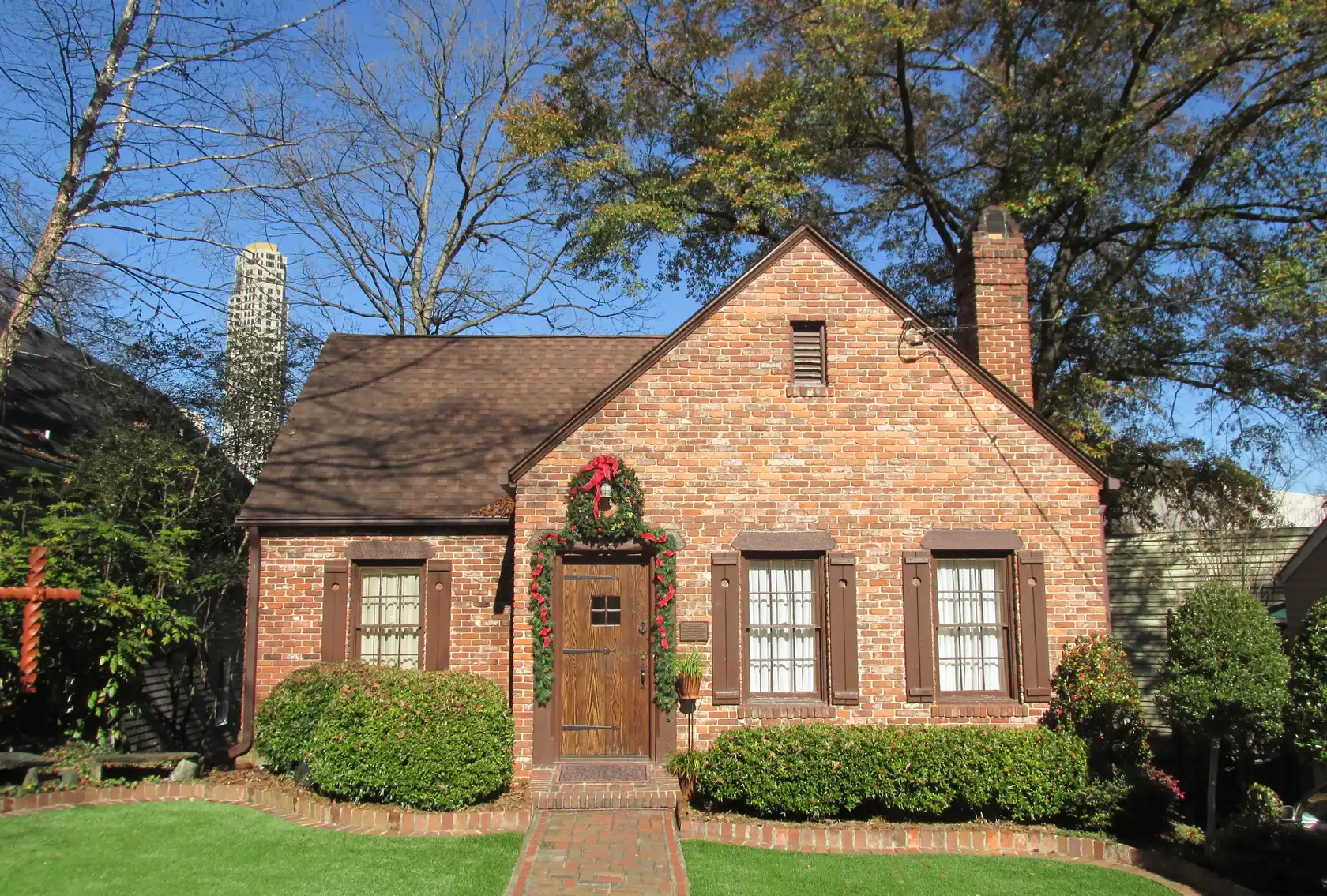 A small red-brick cottage with a steep gable and a wreath on its wooden door, a brick walk across a green lawn, bare trees and a tall cylindrical high-rise above the roofline.