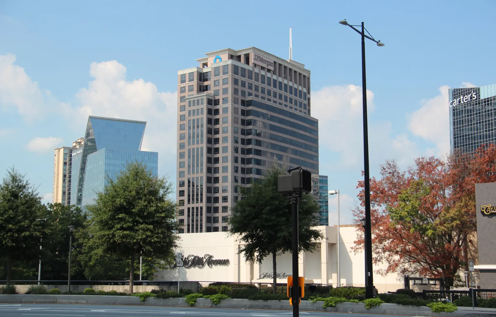 A wide road and planted median at street level, tall office and condo towers behind a low white store building, and a tree with red-orange autumn leaves in front under a blue sky.