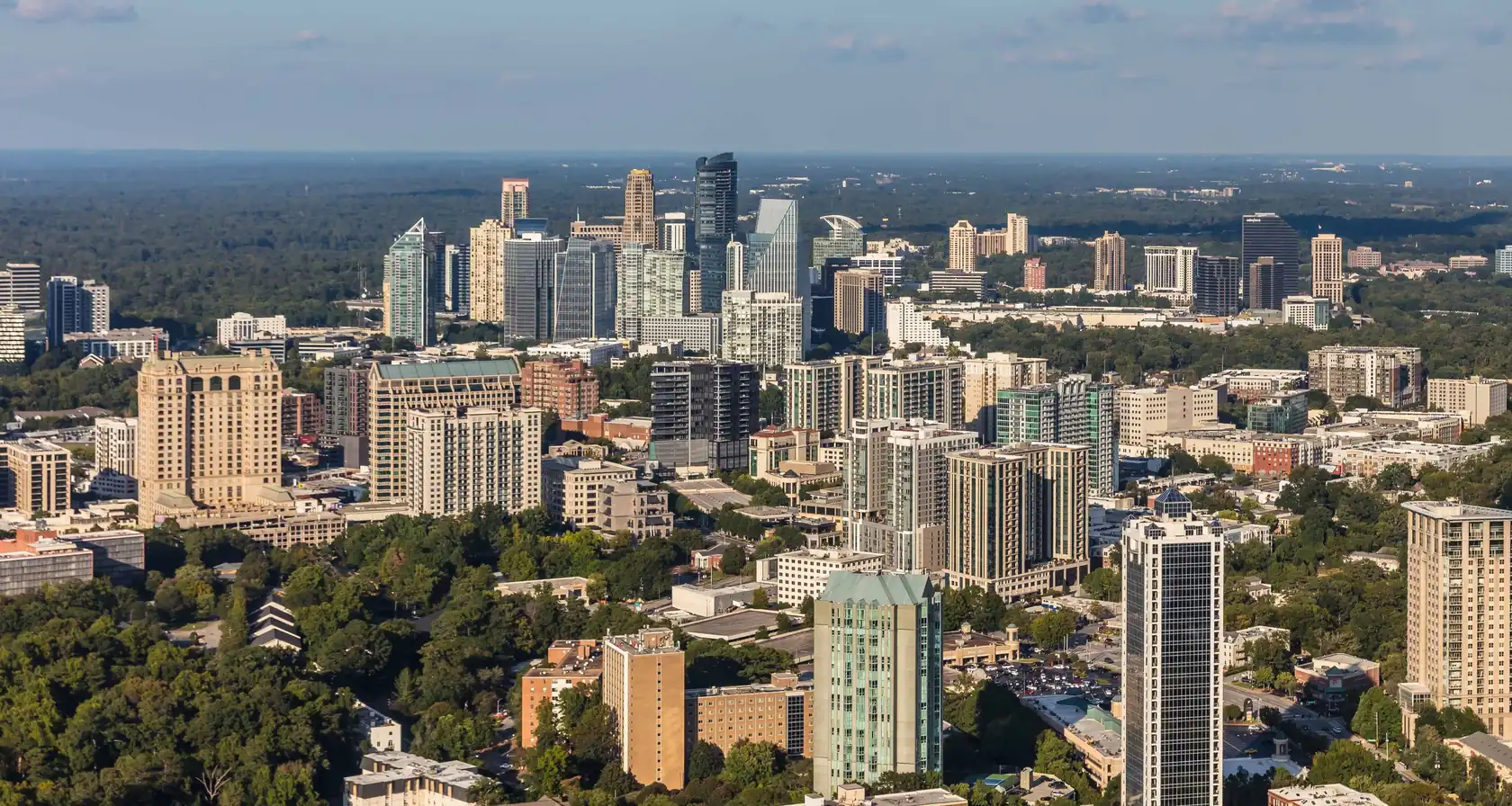 A cluster of glass and stone high-rises of mixed heights rising out of a dense green tree canopy, with forested land running to the horizon under a pale blue sky.