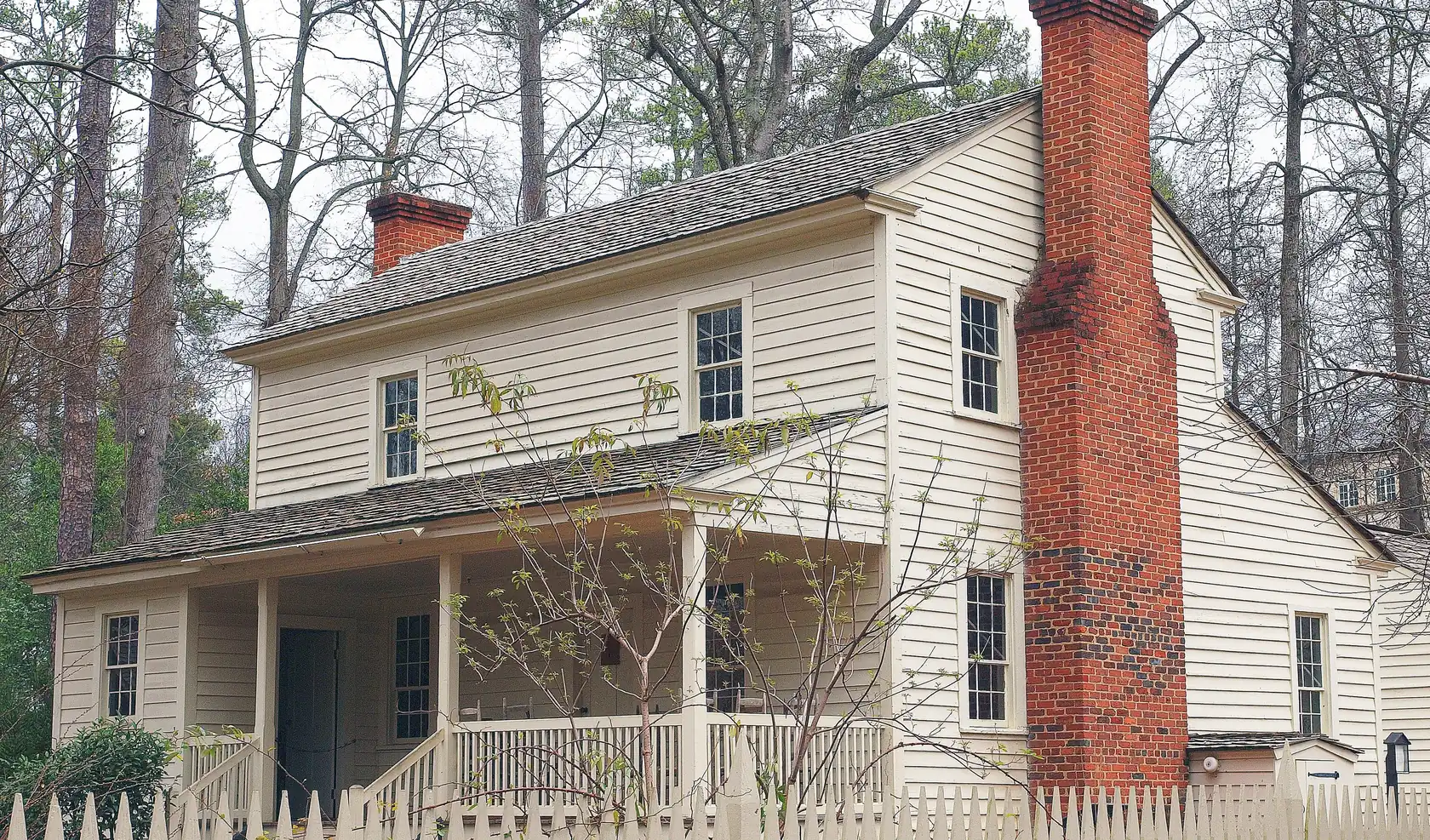 A two-storey white clapboard farmhouse with a long front porch and a massive brick end chimney behind a white picket fence, among tall winter trees.