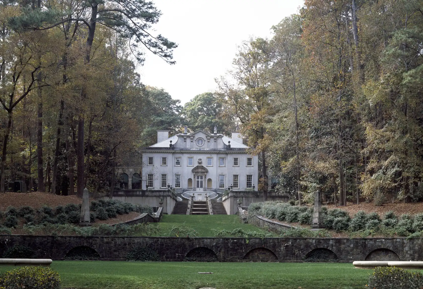 A pale stone mansion at the top of a grass terrace with twin stairs and stone obelisks, surrounded by tall trees turning gold, bronze and rust, with fallen leaves on the slopes.