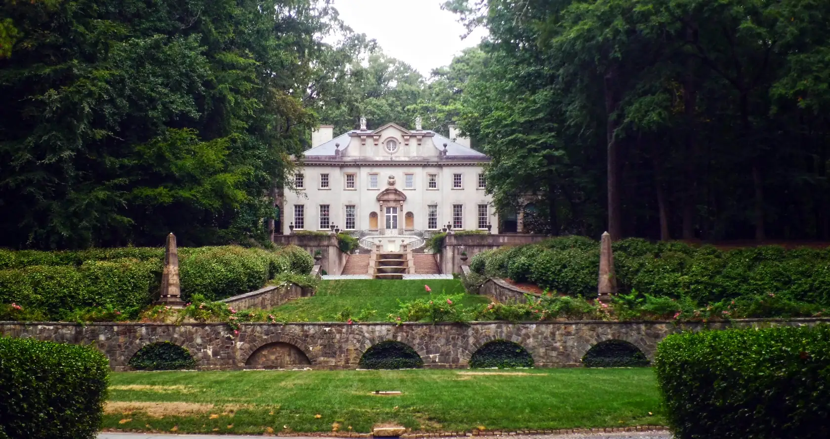 A symmetrical view up a formal terraced garden with an arcaded stone wall, a central water stair and clipped hedges to a pale classical mansion framed by tall dark trees.