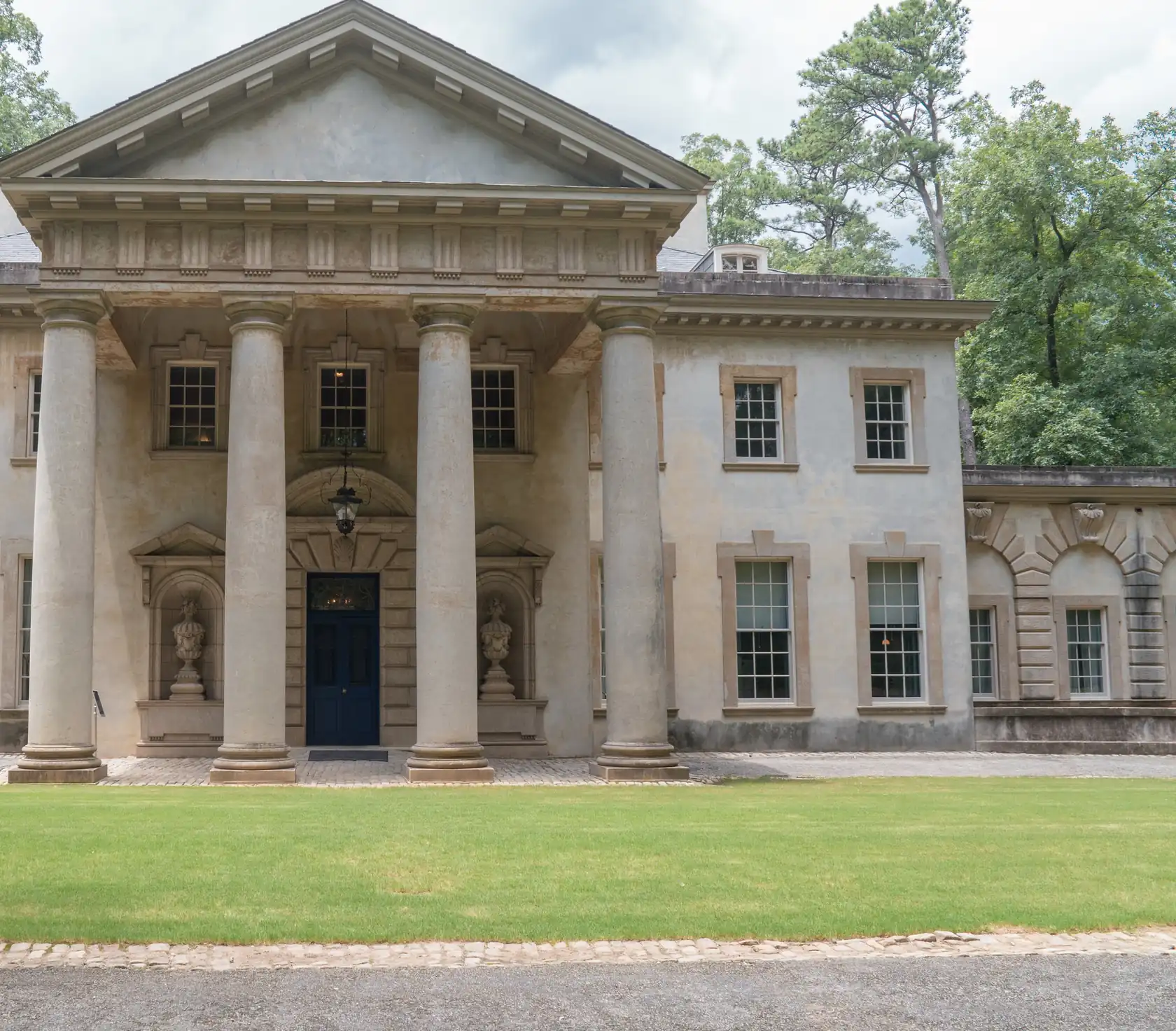 The entrance front of a large pale classical mansion with a portico of four tall columns over a dark blue door, arcaded wings and a broad mown lawn in front.