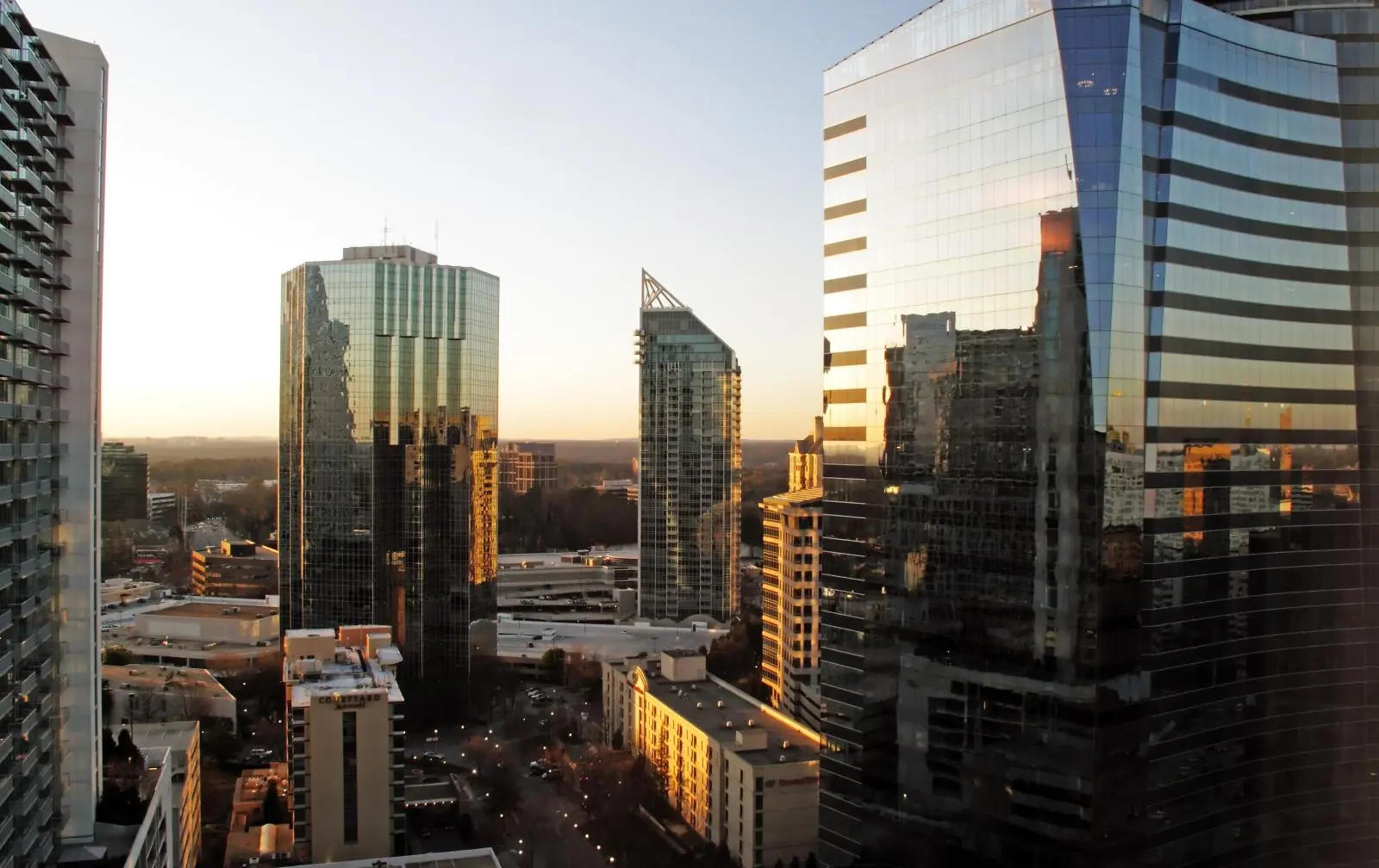 Glass office and condo towers at sunset seen from a high floor, a curved dark-glass tower at right and a slim tower with an open crown at centre, low sun on a street lined with lamps and flat wooded land on the horizon.