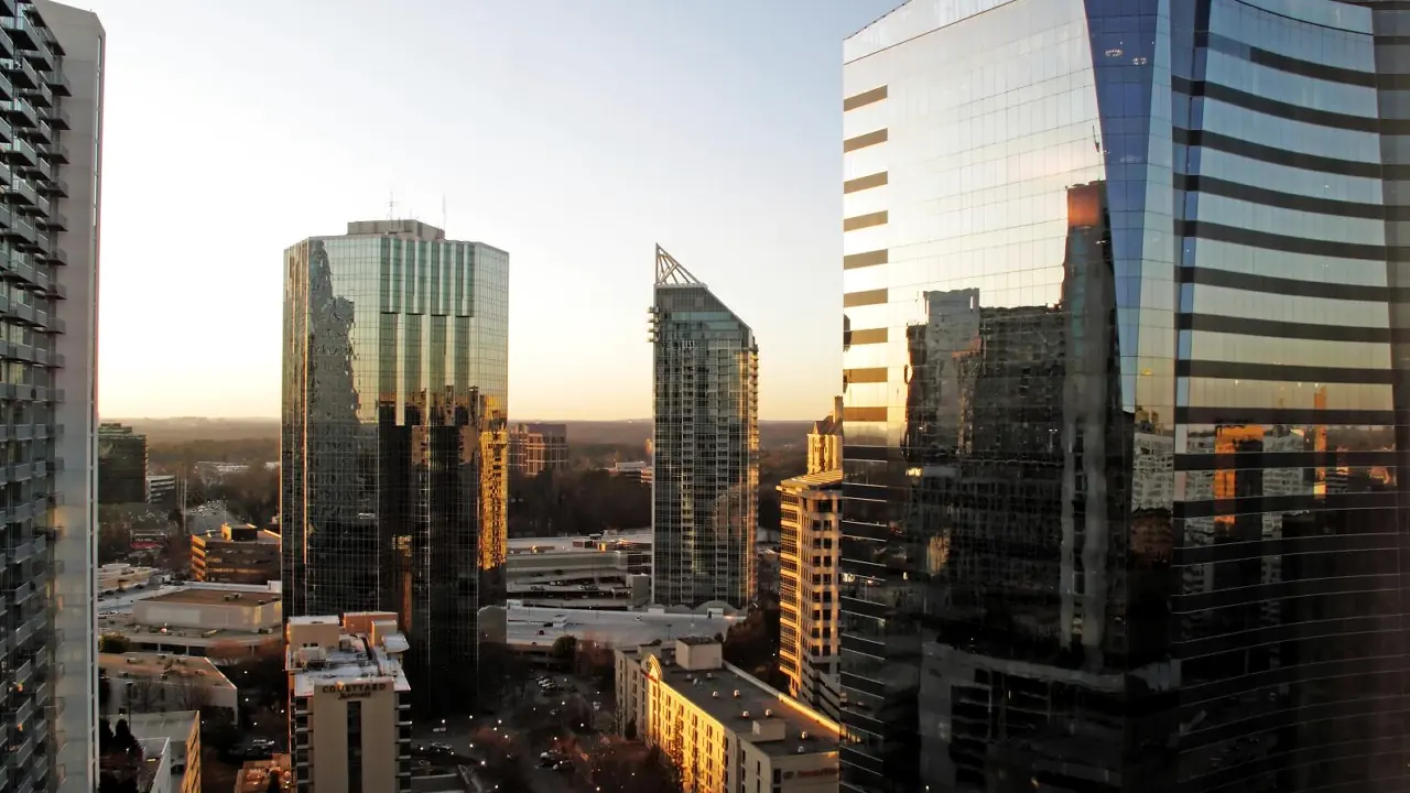Glass office and condo towers at sunset seen from a high floor, a curved dark-glass tower at right and a slim tower with an open crown at centre, low sun on a street lined with lamps and flat wooded land on the horizon.