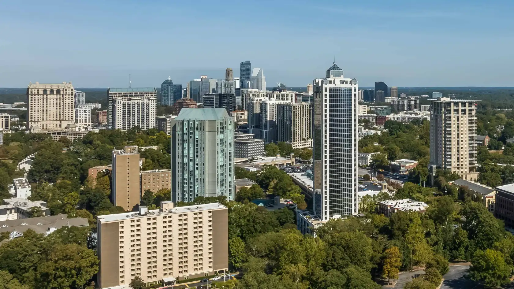 Treetops in front of a group of high-rises: a pale green glass tower, a slim dark-glass tower with a pointed crown and tan stone towers, under a clear blue sky.