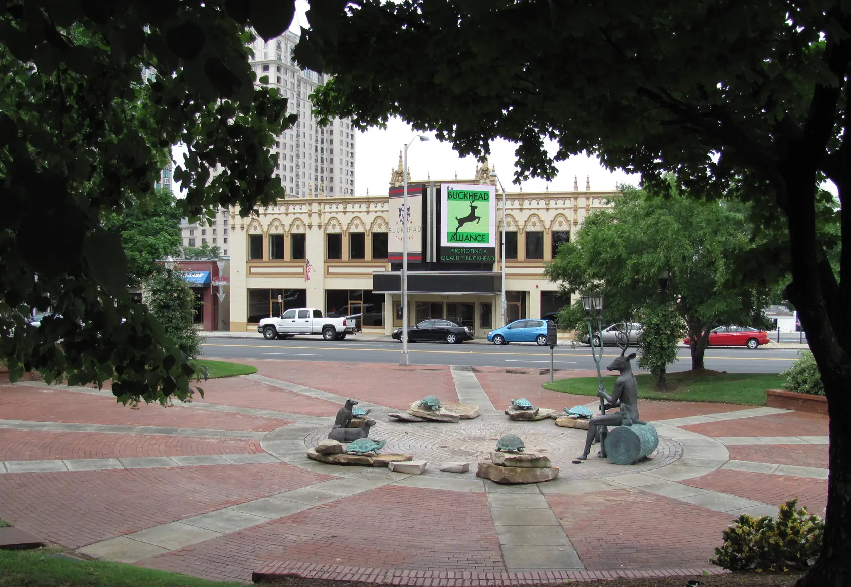 A round brick plaza with bronze sculptures framed by dark branches, a cream two-storey theatre front with ornate pinnacles across the street and a tall beige tower behind.